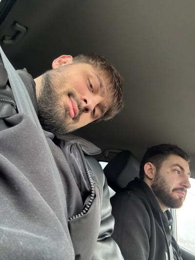 The image depicts the interior of a vehicle, occupied by two adult males. The specific location in the city of Bazan, Bulgaria, cannot be determined from the visual evidence provided.

The primary subject, positioned in the foreground and occupying the left side of the frame, is a male with light brown hair, a beard, and a mustache. He is wearing a black jacket, featuring a visible zipper, and a gray shoulder-belt strap is positioned across his chest. He faces the camera with a tilted head and his tongue slightly extended.

The secondary subject, located to the right and slightly behind the first, is a male with dark hair and a beard. He is wearing a black hooded sweatshirt and is observed in profile, looking towards the right side of the vehicle, out a window. A dark headrest is visible behind him.

The vehicle's interior features a dark headliner with a visible overhead grab handle. The exterior environment visible through the window next to the second subject is indistinct, appearing as a light gray, likely overcast, sky or distant scene.