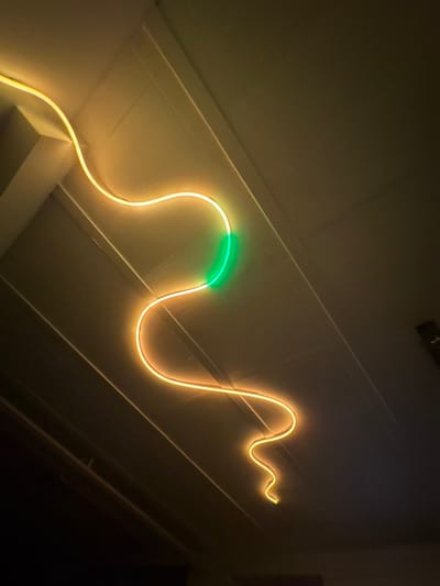 A luminous, serpentine light fixture adorns a white wall. The light emanates a warm, yellow glow except for a segment that transitions to a vibrant green. The fixture's winding path creates an undulating pattern. Its placement accentuates the architectural details of the wall, which include horizontal lines and a corner intersecting the right side of the wall. A dark area is visible at the wall's edge.