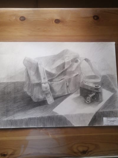 This is a graphite drawing on paper, depicting a still life arrangement. The drawing features a vintage camera placed next to a canvas messenger bag on a surface. The composition is rendered with varying degrees of shading, creating a sense of depth and texture. A small label is affixed to the lower right corner of the drawing, containing text in Cyrillic script. The drawing is presented against a wooden background, likely a tabletop or shelf, with visible wood grain and knots. The overall impression is one of a meticulously executed artistic study. Based on the Cyrillic inscription, the artwork is likely from Yemelyanovo, Russia.