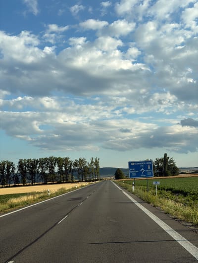 The image shows a straight asphalt road with white lane markings, leading towards the horizon under a sky filled with fluffy white and gray clouds. On the right side of the road, a blue traffic sign is visible, indicating directions to "SLOVENSKE N. MESTO" (Slovenské Nové Mesto), "VEĽKÉ KAPUŠANY 552", and "KOŠICE 552" with a roundabout symbol and distances. The sign also shows "79" which likely refers to a road number. Next to the main sign, a smaller sign indicates "40.0". Fields of yellow and green crops are present on either side of the road, bordered by a line of tall trees in the background. The scene appears to be in a rural area of Slovakia, possibly near Zemplínska Nová Ves, as suggested by the signage. There are no people or vehicles visible in the image.