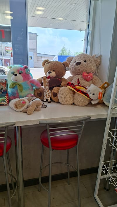The image shows a collection of stuffed animals on a counter, possibly in a shop or cafe, located in Nytva, Russia. The stuffed animals include several teddy bears of various sizes and colors, a giraffe, a snake, and a white plush dog. One large beige teddy bear has a pink heart with "Love You" written on it. Another large teddy bear is wearing a yellow and red striped hat. A tie-dye colored teddy bear is on the left side of the counter. There are also two red bar stools in the foreground, suggesting the area may be a seating area. In the background, through a window, a building and trees are visible. A sign with Russian text that reads "ЛОМ" is visible on the building. The overall scene is indoors with natural light coming from the window.