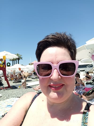 A close-up, front-facing selfie shows a fair-skinned woman with dark hair and pink-framed sunglasses. She is smiling, her lips painted with coral lipstick. She is wearing a dark bikini with a colorful, abstract pattern. The background is a sunny beach with numerous people relaxing on lounge chairs under white umbrellas. Palm trees are visible in the distance, suggesting a warm climate. The beach appears to be covered in grey pebbles. A timestamp, "2026/6/21 13:07," is visible in the lower right corner, implying the image was taken on June 21, 2026, at 1:07 PM. Based on the palm trees and beach setting, this is likely a location in the city of Bolshoy Sochi, Russia.