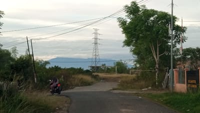 A person rides a dark-colored motorcycle on a paved road that curves to the left. The rider, facing away from the camera, wears a striped long-sleeved shirt and dark pants. The motorcycle has a visible rear brake light. The road is flanked by overgrown grass and vegetation on the left and a dirt area with scattered debris on the right. In the background, a tall metal electricity pylon stands prominent against a cloudy sky. Further in the distance, a mountain range is visible, partially obscured by trees and haze. Two soccer goals are positioned on a field in the mid-ground. To the right of the frame, a fenced property with an orange-colored wall and a dark sign with white text is present. The scene appears to be in Meunasahteungkudigadong, Indonesia, given the context.
