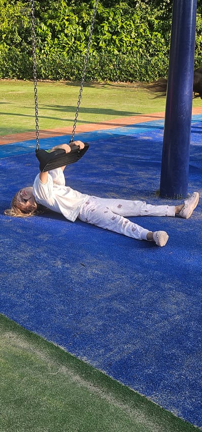 A young girl with blonde hair, wearing white pajamas and pink clogs, lies on her back on a blue rubber playground surface, holding onto a black swing seat. The swing is suspended by chains from above. The playground is bordered by green turf. In the background, a dense green hedge forms a backdrop. A large blue pole is visible on the right side of the image. The scene is set outdoors during the day, likely at a playground in Groenlo, Netherlands, given the context.