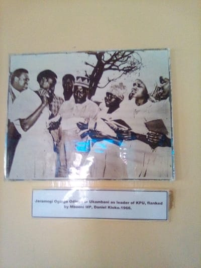 This image displays a framed, black and white historical photograph mounted on a light beige or yellow wall, observed within a building located in the city of South Sakwa ward, Kenya. Below the main photograph, a white rectangular caption label with black text is affixed to the wall.
The photograph depicts a group of eight individuals, primarily men, gathered outdoors under a tree with visible, sparse branches against a light background. The central figure, identified by the caption as Jaramogi Oginga Odinga, wears a light-colored long-sleeved shirt and a distinctive, patterned cap. He is seen holding a small object or document in his hands. To his immediate right, another man, identified as Mbooni MP Daniel Kloko, is partially visible. To Oginga Odinga's left, a man in a light shirt is actively speaking, gesturing with his left hand towards Odinga. Another individual to Odinga's far right, wearing a patterned head covering, holds papers or a book. Other figures are visible in the background, some partially obscured. Several individuals in the photograph are holding papers, documents, or small books. The scene appears to be an outdoor gathering or meeting.
The caption below the photograph reads: "Jaramogi Oginga Odinga in Ukambani as leader of KPU, flanked by Mbooni MP, Daniel Kloko. 1966." The photograph itself is covered by a reflective surface, possibly glass or plastic, which shows some glare, particularly in the lower right quadrant. The frame enclosing the photograph has a thin, dark border.