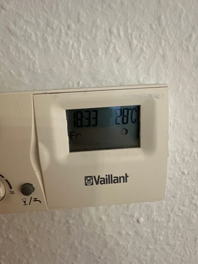 The image shows a close-up of a Vaillant brand digital thermostat mounted on a textured white wall, likely in a home in Recklinghausen, Germany. The thermostat display shows the time as 18:33 and the day as "Fr" (Friday). The current temperature reading is 28°C, indicated by a small circle symbol below the temperature. To the left of the display, there is a circular dial with a pointer set at "30" and a dark gray button. Below these controls, there is a symbol that appears to be a wine glass next to a water tap. The wall's texture is rough and uneven, typical of many interior walls. No people are visible in the image.