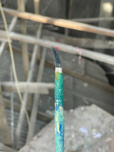 This close-up image portrays a single, well-used paintbrush positioned vertically in the foreground, sharply focused. The brush has dark grey/black bristles, which are splayed and appear worn, with a distinct small red speck visible near their tip. A silver-colored metallic ferrule connects the bristles to the handle. The handle is primarily teal/green, heavily textured and caked with dried splatters of various paint colors, including yellow, blue, and white. The background is significantly blurred, depicting a structure composed of light-colored wooden or bamboo poles arranged in a crisscross, scaffolding-like pattern. A thin yellow wire or rope is faintly visible on the left side of the blurred background, and a small red and white striped object is discernible on one of the background poles. The environment appears to be an outdoor setting, possibly a construction site or workshop. No individuals or active interactions are present in the scene. The location is in the city of Mns Asan, Indonesia.
