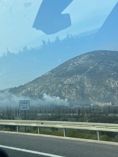 The image captures an outdoor scene in Kırkağaç, Turkey, viewed from inside a vehicle. The foreground features a multi-lane paved road with a white solid line on the left and a white dashed line on the right, bordered by a metal crash barrier. Beyond the barrier, a strip of green vegetation, including bushes and a line of slender trees, precedes a large, rocky mountain that dominates the background. A prominent plume of light gray smoke emanates from an area at the mountain's base, indicating a fire or controlled burn. This smoke partially obscures distant buildings and utility structures located in the midground. A white rectangular sign with black text, displaying "0-5/21" on the top line and "002" on the bottom line, is visible in the mid-left of the frame. The sky is light blue, transitioning to a hazier tone near the horizon. Reflections from the vehicle's interior are present in the upper portion of the image. No individuals are visible.