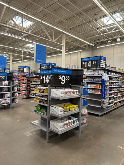 The image displays an interior view of a retail store, likely a large discount chain such as Walmart, located in Worcester, United States. The perspective is tilted, showcasing several aisles lined with shelves of products. The ceiling features exposed white trusses, ductwork, and numerous fluorescent lights, along with visible skylights, indicative of a warehouse-style building.

In the foreground, grey metal shelving units hold various packaged goods, many of which appear to be wellness, health, and personal care items. Prominent black and white signs are attached to the shelves, advertising "Everyday Low Price" with specific prices such as "$14.97", "$9.88", and "$14.4?", corresponding to aisle sections like "G7", "G9", "G11", and "G13". A blue hanging sign above the aisles clearly labels a section as "Wellness." Further down the aisle, a product display includes text in both English and Spanish: "Bienestar para la Familia" and "Family Wellness." The store floor is a simple, polished concrete. No people are visible in the frame, presenting a static view of a typical retail environment during what appears to be regular operating hours.