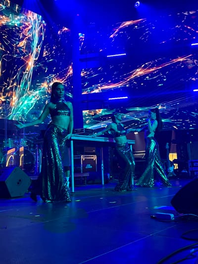 The image depicts a stage performance with three women in matching sparkling green outfits. The woman on the left is in the foreground, facing the audience and extending her left arm outwards. Two other women are positioned behind her, closer to a table with electronic equipment. The stage is lit with vibrant blue and abstract colorful lights, suggesting a concert or performance setting. The backdrop features a dynamic projection of abstract, glowing lines and shapes. In the background, drum equipment and other stage apparatus are visible. The flooring appears to be a dark, reflective surface. There is no visible information in the image to suggest the location is the city of Akhalubani, Georgia.