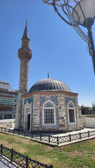 In İzmir, Turkey, a stone minaret and the domed structure of a small mosque stand against a clear blue sky. The mosque's exterior is adorned with intricate blue and white tilework framing arched windows. A low, decorative black metal fence surrounds the mosque and a patch of green grass. In the background, modern buildings rise, contrasting with the historic mosque. A street lamp with ornate lights is visible in the upper right corner. There are no people or animals present in the image.