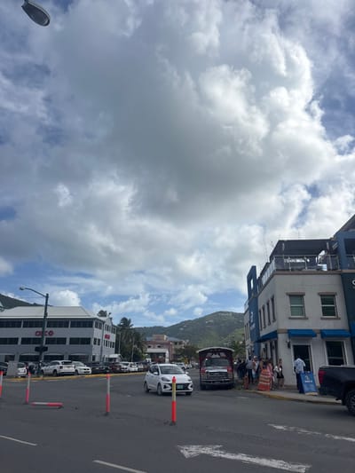 An outdoor street-level scene in Road Town, British Virgin Islands, is depicted under an overcast sky dominated by large cumulus clouds with patches of blue. The image captures a paved road in the foreground with a white left-turn arrow painted on the asphalt. Several red and white striped flexible bollards are present along the road, one of which is lying horizontally.

On the left, a multi-story building with a grey exterior features "CIBC" branding on its facade, behind a streetlamp with a dark green pole. A parking lot in front of this building contains multiple parked vehicles. Towards the center of the road, a white compact five-door hatchback is in motion, approaching the foreground.

On the right side of the image, a two-story commercial building displays a light blue and white facade with blue window awnings on the lower level. A rooftop terrace with seating and glass railings is visible. Positioned along the curb in front of this building is a red open-air tourist shuttle vehicle, resembling a modified truck, with its hood slightly ajar. A group of approximately ten to fifteen individuals, appearing to be pedestrians or tourists, are gathered on the sidewalk in front of the building and near the shuttle. One person in a light blue shirt stands next to an A-frame sign. Part of a dark-colored pickup truck is visible on the far right.

In the mid-ground, other buildings, including a light pink structure, are visible. The background features lush green, undeveloped hills and mountains under the cloudy sky.