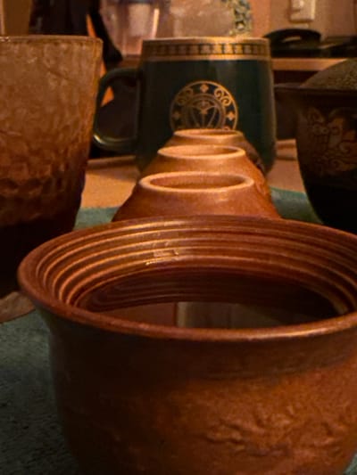 The image shows a close-up, Dutch angle shot of several ceramic items, possibly mugs or vases, in a warm, indoor setting. The primary subject is a large, reddish-brown ceramic vessel with a ribbed interior, taking up the right side of the frame. To its left, a dark green ceramic mug with a decorative gold band and emblem is visible. The background is indistinct, with hints of other ceramic objects and a textured surface that might be fabric or another ceramic piece. The lighting appears artificial, suggesting it's likely indoors, and the overall tone is cozy and rustic. There is no visible text in the image. The scene does not depict any people or specific events, focusing solely on the decorative qualities of the ceramic objects. The context of Pevek, Russia, does not offer direct visual cues within the image itself, but implies a potentially cold climate outside, which might contribute to the cozy indoor atmosphere.