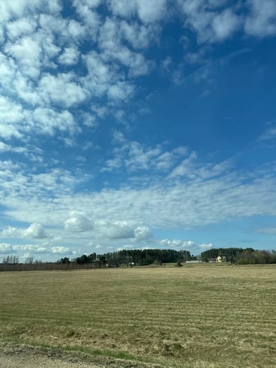 The image shows a field in Ādaži, Latvia, partially covered in dry grass. A line of trees separates the field from a sky filled with scattered, puffy clouds and patches of blue. A yellow building is visible in the distance beyond the trees. The photo is rotated.