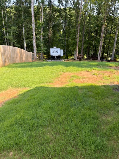 The image shows a camping trailer parked among trees. The trailer is white with "BLACK LABEL" written on the back. A wooden deck is visible in the foreground. The scene is an outdoor setting with a lawn and trees. The lighting suggests it is daytime, likely late afternoon, with a shadow cast across the lawn from the trees. The overall impression is a peaceful, natural environment. The image is taken from a slightly elevated angle, looking down and across the scene.