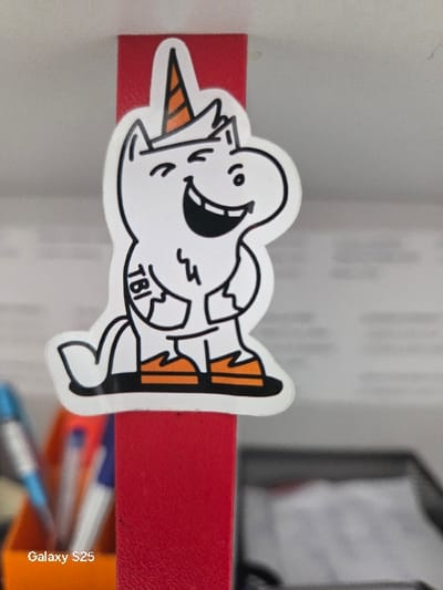 A white unicorn sticker with a black outline, an orange-striped horn, and orange feet is affixed to a textured, vertical red surface in an indoor environment. The unicorn character has a winking left eye, a smiling right eye, and an open mouth with visible teeth. The letters "TBI" are present on its left hip. In the blurred background to the lower-left, an orange desk organizer contains two pens, one blue with a silver tip and one entirely blue. To the lower-right, a dark container holds white papers. The scene is located in Novo Delchevo, Bulgaria, depicting an office or workspace.