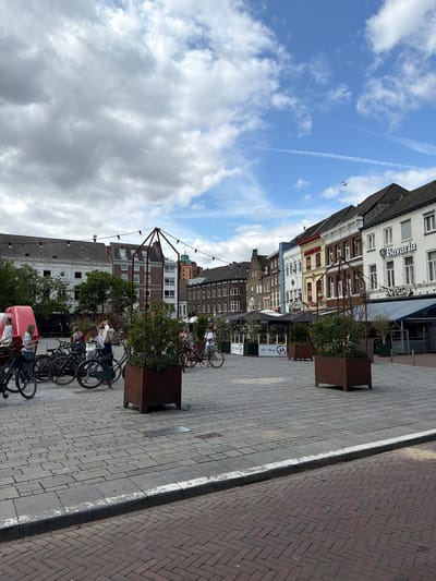 The image depicts a street scene in Roermond, Netherlands. In the foreground, the ground is paved with interlocking grey stone bricks, with a distinct curb lined with white paint. To the right, a herringbone pattern of dark red bricks is visible. In the middle ground, several people are gathered around bicycles. A group of four women, some of whom are holding phones, stand near a red, inflatable-like structure. To the right of the women, a row of parked bicycles is visible. Further back, in the background, a row of European-style buildings line the street. One building prominently displays the "Bavaria" logo. String lights are strung across the street between poles. Two large, square planters with greenery are placed on the paved area. The sky above is partly cloudy, with patches of blue sky visible.