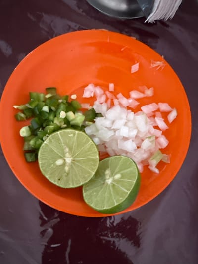 An orange plate is placed on a dark, reflective surface. The plate contains chopped green jalapeños, diced white onions, and two lime halves. The jalapeños are finely diced and mixed with some seeds. The onions are also finely diced and appear translucent in places. One lime half is positioned to the left of the plate, showcasing its radial segments. The other lime half is to the right and slightly below the first, also showing its internal structure. The background is dark and shows some subtle reflections, suggesting it might be a table. A sliver of a metallic object and a stack of white napkins are visible in the upper right corner of the image. The scene appears to be set in Tizapan, Mexico, likely a casual dining or food preparation setting, given the ingredients and presentation.