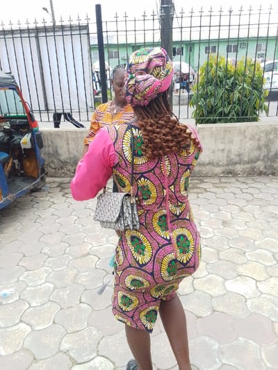 The image shows two women walking on a paved street in Yenagoa, Nigeria (4.931, 6.268). The woman in the foreground is wearing a pink and yellow patterned dress with a black purse slung over her shoulder. She has her hair braided and is wearing a pink head scarf. The woman in the background is wearing a yellow and brown patterned dress. There is a black auto rickshaw parked in the background. The scene takes place in a street with an iron fence in the background. The iron fence separates the street from a green building in the background. The women are walking towards the right side of the image. The woman in the foreground is looking over her shoulder to the right. The woman in the background is looking at the woman in the foreground. There is a lot of greenery in the background, including some palm trees.