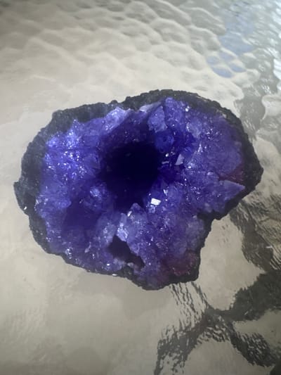 A close-up, high-angle shot shows a dark, rough exterior geode, filled with sparkling, crystalline amethyst, resting on a reflective, textured surface. The geode's interior is a vibrant purple, with sharp, multifaceted crystals catching the light. The exterior is a dark, matte black, with an irregular, rocky texture. The surface beneath the geode appears to be glass or a similar material with a patterned texture, causing reflections of the geode and ambient light. The image is undated and unlocated within Delray Beach, United States of America.