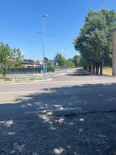 This image captures a street scene in Castiglione delle Stiviere, Italy, on a clear, sunny day. The foreground is dominated by an asphalt road, with shadows from nearby trees dappling its surface. In the mid-ground, a paved intersection is visible, featuring a pedestrian crossing and several streetlights. Traffic signs are present, including a "No Stopping" sign, a "3.5t" weight limit sign, and signs indicating bicycle paths and a one-way street. A row of trees lines the right side of the road, leading into the distance. On the left side of the street, houses with tiled roofs and green fencing are visible. The sky is a vibrant blue, with a few wispy clouds. The overall impression is of a quiet, well-maintained urban or suburban environment.