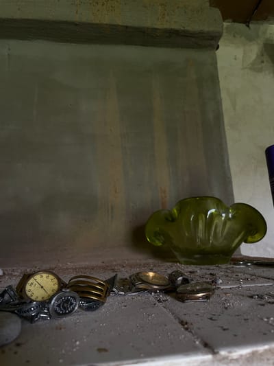 The image depicts an interior scene, likely within a building. The foreground is cluttered with several wristwatches, some appearing vintage, scattered across a dusty, textured surface that resembles a shelf or countertop. The watches vary in design, with visible faces and bands. Among the watches are other small metal objects, possibly jewelry or accessories, also showing signs of age and disuse.
In the midground, to the right of the watches, sits a vibrant green glass ashtray or bowl with a distinctive, ruffled rim. Its shape is organic and reminiscent of a flower. Behind the ashtray, the wall is rendered in a muted, light gray with vertical streaks of yellow and reddish-brown, suggesting possible water damage or staining.
The background consists of a concrete structure, possibly a fireplace or architectural element, with a rough, textured surface. Above this, a dark wooden beam is visible, indicating the ceiling or upper structure of the room. The overall lighting is dim, casting subtle shadows and highlighting the dusty, neglected state of the objects and the environment. The scene evokes a sense of abandonment or disarray.