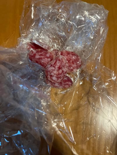 The close-up image shows several slices of salami wrapped in clear plastic cling film, resting on a light brown wooden surface, likely a table or countertop, in Chêne-Bourg, Switzerland. The salami slices are round and display a reddish-pink color with distinct white specks of fat distributed throughout. A fine, powdery white coating, characteristic of some cured meats, is visible on their surfaces, particularly around the edges. One slice is partially visible, showing its thin profile. The transparent plastic wrap is crinkled and reflects light from an unseen source, creating bright highlights and subtle shadows across its folds. No people or specific activities are depicted, suggesting the salami is either stored, being prepared, or awaiting consumption. No text is visible within the frame.