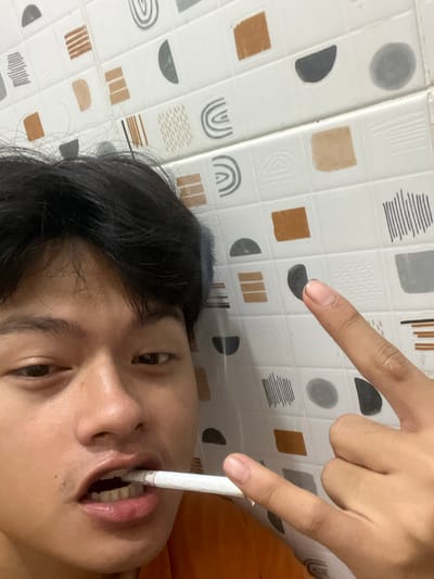 A close-up, low-angle, point-of-view shot captures a young individual, seemingly male, with dark hair and brown eyes, holding a white object, possibly a cigarette or vape, in their mouth. The individual's head is tilted, and their mouth is slightly open, revealing teeth. One hand is visible, with fingers forming a rock-and-roll gesture and another finger pointing towards the background. The background consists of a tiled wall with a repetitive pattern of abstract shapes in brown, grey, and white. The scene appears to be indoors, possibly a bathroom, given the tiled surface. The location is identified as Serdang Kulon, Indonesia.
