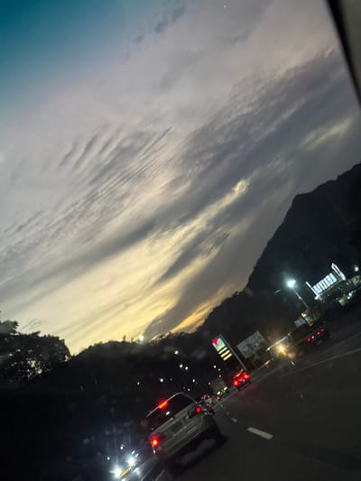 The image shows a view from a vehicle on a highway at dusk or night. The sky is filled with striated clouds, ranging in color from light gray to yellow. On the right side of the frame, the highway is visible with cars and their headlights and taillights illuminating the road. Some buildings are illuminated on the side of the highway. The location is likely near a city, though the name Cortada de Maturín, Venezuela, cannot be definitively determined from this image alone.