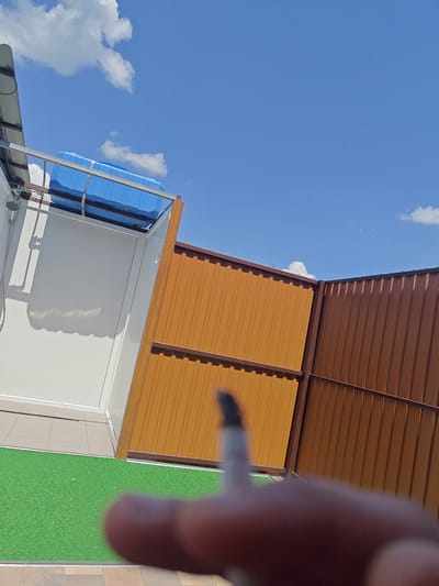 In Shkurupiivka, Ukraine, a scene unfolds under a bright blue sky with scattered clouds. In the foreground, a person's hand holding a lit cigarette is blurred. Behind it, a section of artificial green turf is visible, adjacent to a paved area. Further back, a light brown corrugated metal fence meets a darker brown corrugated metal fence, forming a boundary. To the left of the fence, a white outdoor shower structure stands, topped with a blue water tank. The shower stall has a visible shower head and piping.