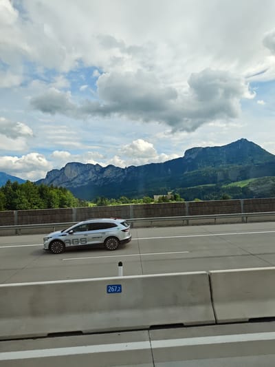 The image shows a highway scene in St. Lorenz, Austria, with a white SUV driving on the right lane. The SUV has "AGS" and "www.ags.at" written on its side. In the background, a range of mountains covered in lush green trees rises against a sky with dramatic clouds. A wooden fence separates the highway from the trees. The foreground features concrete barriers with a blue sign that reads "267,2". The scene is captured during the daytime under a cloudy sky.