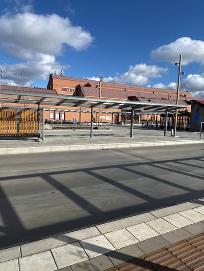 The image depicts an urban landscape in Jönköping, Sweden, featuring a street with a building facade on the left. The building is characterized by its reddish-brown exterior, windows with red frames, and a complex roof structure. A steel framework runs along the building, casting shadows on the pavement. The street surface is asphalt with a concrete curb. Tactile paving is visible adjacent to the curb. A bus stop with seating is present. The sky is blue with cumulus clouds.
