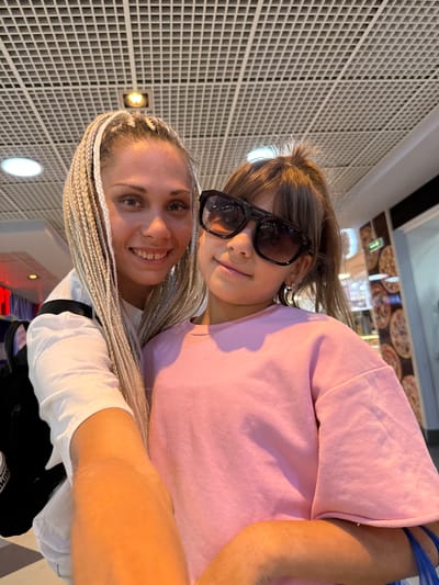 In an indoor setting, possibly a mall or food court, a woman with blonde braided hair and a young girl wearing sunglasses are posing for a selfie. The woman is wearing a white top and a black backpack. The girl is wearing a pink sweatshirt. Behind them, the ceiling features a grid-like structure with lights. To the right, there is a display or wall featuring images of pizzas. The location is identified as Serenovo, Russia.