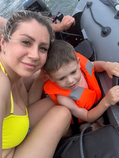 The image shows two people, a woman and a young child, on a boat. The woman is in the foreground, wearing a yellow bikini top. The child is wearing an orange life vest with gray reflective strips. The boat appears to be an inflatable dinghy with gray tubes and black seats. The water is visible in the background, suggesting the boat is on a lake or river. The overall mood seems relaxed and happy, with both subjects appearing to be enjoying themselves. The time of day appears to be daytime, and the weather seems to be pleasant, conducive to being on a boat. There is a small outboard motor visible at the back of the boat. The woman is looking at the camera, while the child is looking up and smiling. There is no visible text in the image. The context provided places this scene in Pravda, Russia.