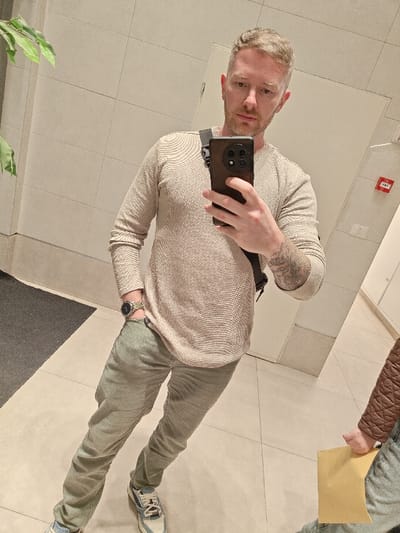 A man stands in a tiled room, holding a phone to take a selfie. He wears a beige long-sleeved shirt, gray pants, and patterned sneakers. A black bag strap crosses his body. He has short blonde hair and a light beard. A watch is visible on his left wrist. Part of a green plant is in the background on the left. Tiles cover the walls and floor, with a section of dark gray flooring also visible. Another person, partially visible on the right, holds a yellow paper.