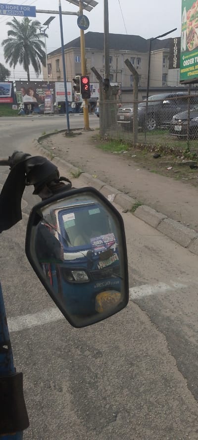 The image displays an urban street scene in Warri South, Nigeria, captured from the viewpoint of a vehicle, evidenced by its left rearview mirror occupying the lower left foreground. The mirror reflects the front of a blue tricycle (Keke Napep), revealing a driver, a partially visible license plate "1128-RT," and a "JESUS CHRIST" poster on the windshield, along with the reversed text "adesarot" on the vehicle's front.

The immediate ground consists of an asphalt road surface with a white painted line, transitioning to a concrete curb and a dirt shoulder on the right. In the midground, a traffic intersection is visible. A yellow pole supports a solar panel array, a blue pedestrian crossing sign, and a traffic light displaying a red signal with a numerical countdown of "2." Adjacent to this, a chain-link fence encloses an area with several dark-colored passenger vehicles parked within.

To the left of the intersection, an overhead blue sign reads "...D HOPE FOR ...LE AGENDA." A prominent palm tree stands near multiple large advertising billboards, one partially displaying "ONE WAY YOU CAN CHANGE YOUR..." and another advertising pizza with "ORDER TO..." Several other blue tricycles and indistinct human figures are present further down the street near these billboards. The background features a multi-story building with light-colored walls and a dark roof, identified as "PRINCIPALE HOTEL," alongside another similar structure. The sky is overcast.