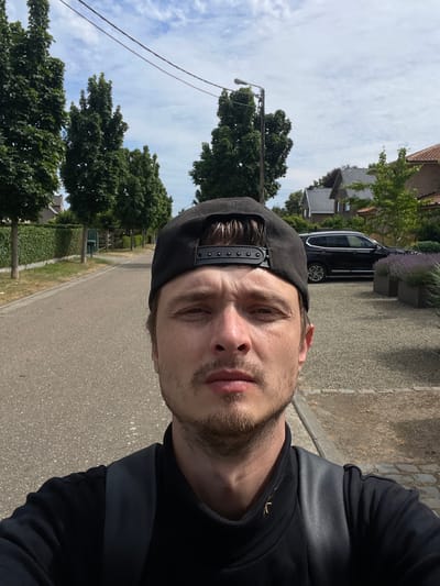 The image is a selfie taken outdoors on a street in Diepenbeek, Belgium. The subject, a man wearing a black baseball cap and a black shirt, is positioned in the foreground, facing the camera. Behind him, a paved road runs parallel to a neatly trimmed green hedge on the left side and a gravel driveway with lavender planters and houses on the right. A dark colored SUV is parked on the driveway. The sky is partly cloudy, and power lines are visible overhead, stretching across the frame. The scene suggests a residential area with well-maintained properties and a calm, daytime atmosphere.