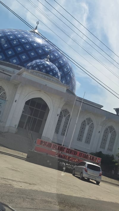 The image shows the Masjid Islamic Centre At-Taqarrub in Paya Tringgadeng, Aceh, Indonesia. The mosque features a large blue dome with a white geometric pattern. A silver car is parked in front of the mosque on the road. A sign reading "MASJID ISLAMIC CENTRE AT-TAQARRUB, KUALA IDIE, ACEH TIMUR, PAYA TRIENGGADENG" is visible. Overhead power lines are present in the top left of the frame. The sky is blue with scattered clouds.