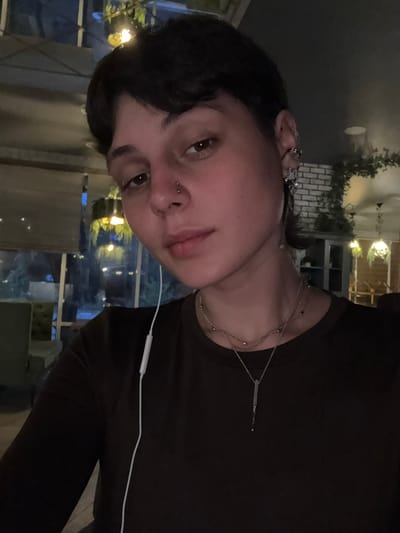A close-up, medium shot of a person with dark hair and fair skin, facing the camera with a slight tilt. The person has a nose piercing and is wearing multiple delicate silver necklaces and ear jewelry. They are wearing a dark-colored shirt or top. A white earphone cord is visible across their chest. The background shows a blurry view of what appears to be an outdoor or semi-outdoor dining area at night, with lights and greenery visible. The location in the city of Uryupinsk, Russia, is not discernible from the image.