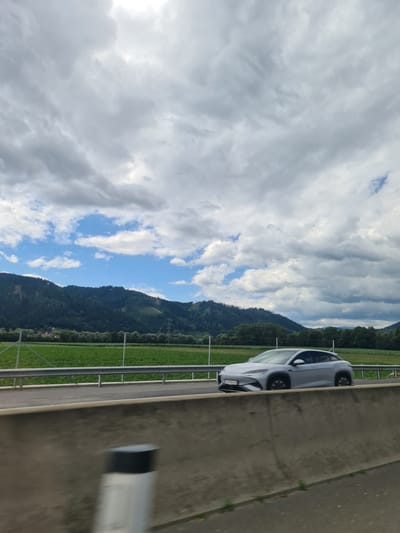 A silver electric car is traveling on a highway in St. Stefan ob Leoben, Austria. The car is in motion and is captured in a side profile. In the background, there are green fields and a dense forest covering rolling hills. The sky is partly cloudy with patches of blue sky visible through the clouds. A concrete barrier is in the foreground, partially obscuring a reflective post.