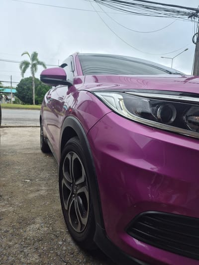 An eye-level, close-up, outdoor, vertical shot shows the front passenger side of a purple Honda HR-V parked on a gravel surface. The car's shiny paint reflects the overcast sky. A portion of the front fender, wheel, headlight, and side mirror are visible. The wheel is a black alloy with silver accents. In the background, a paved road runs parallel to the car, with a thin patch of green grass separating the gravel and the road. Beyond the road, a few trees and buildings can be seen under a cloudy sky. Overhead, a complex network of power lines stretches across the frame, supported by wooden poles. The scene appears to be in a semi-urban or suburban area of Ban Yid, Thailand. There are no people or animals in the image, and no actions are taking place.