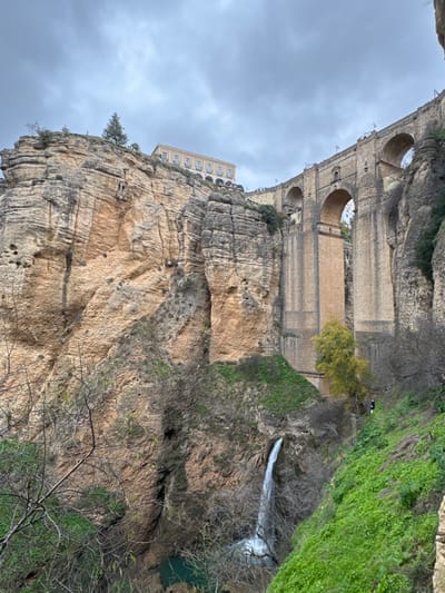 The image depicts the El Tajo gorge in Ronda, Spain, viewed from below and to the left. The prominent subject is the Puente Nuevo bridge, a large stone structure spanning the deep gorge with multiple arches and a tall central support. The bridge features stone balustrades and lampposts along its upper edge, where several small figures of individuals are discernible.

To the left of the bridge, a towering, multi-layered rock cliff rises steeply, displaying striations of tan, grey, and brown rock. On top of this cliff, a light-colored, multi-story building with numerous arched windows is visible, along with a solitary coniferous tree.

At the base of the central bridge support, a significant waterfall cascades into a small, vibrant turquoise-colored pool situated at the bottom of the gorge. Bare-branched trees and sparse green vegetation cling to the lower sections of the cliffs and foreground. A single tree with yellow-green foliage stands on a ledge near the waterfall's origin.

On the right side of the gorge, a steep, vegetated slope descends, revealing a visible dirt path. A single individual, dressed in dark clothing with a prominent white accent, is observed on this path, appearing to be in motion. The sky is overcast with dense, grey clouds, casting diffused light across the scene.