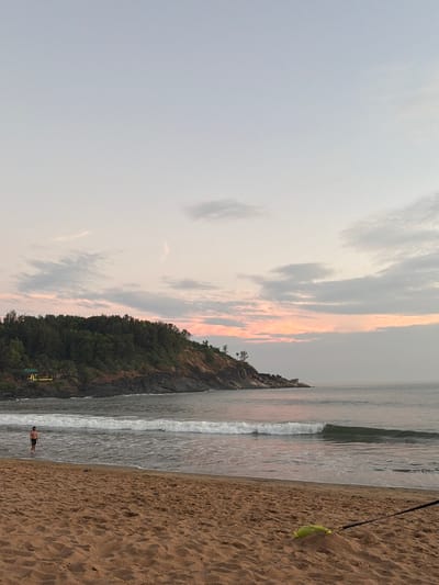 This image depicts a wide-angle view of a beach in Gokarna, India, likely Kudle Beach, during either sunrise or sunset.

The scene is composed of a sandy beach in the foreground, leading into the ocean with breaking waves, and a prominent, tree-covered rocky headland extending into the sea on the left.

The sky, occupying the upper two-thirds of the frame, transitions from a pale blue-grey overhead to soft pink and orange hues near the horizon, characteristic of dawn or dusk. Scattered light grey clouds are present, with some reflecting the ambient pink light.

The ocean's surface is relatively calm further out, while closer to shore, white-crested waves are observed breaking. The water reflects the subtle colors of the sky.

On the left, a dark rocky headland rises from the water, its upper slopes densely covered with green trees and vegetation. A small, light-colored structure with a green roof is partially visible nestled at the base of the headland amidst the foliage.

The sandy beach in the foreground is light brown, marked with footprints and areas wet from receding waves. A single individual, dressed in dark shorts or swimwear, stands waist-deep in the surf in the mid-left portion of the frame, oriented slightly to the right. In the lower right corner of the beach, a lime green object, possibly a bag or tarp, is partially buried in the sand, with a black strap extending from it towards the right edge of the image.