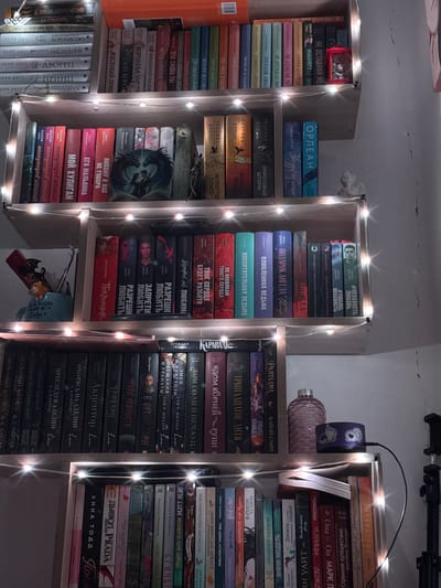 This media file depicts a dimly lit room, likely a bedroom or study, featuring several bookshelves filled with books. The shelves are adorned with strands of small, warm-toned fairy lights, casting a cozy and atmospheric glow. The books are arranged in vertical stacks, with some spines facing outwards, revealing titles and authors. Many of the books appear to be novels, with titles in Russian, such as "Ненависть Любовь", "Подарок Ангела", and "Мой Хулиган".

On one shelf, a decorative, ribbed cylindrical object sits next to a small, purple item with a cartoon character on it, possibly a speaker or a night light. Another shelf holds a small, stylized book cover featuring a dark, bird-like silhouette. The overall impression is one of a personal collection of literature, arranged with a decorative touch. The lighting suggests it is either nighttime or the room is intentionally kept dim. The setting seems domestic and personal.