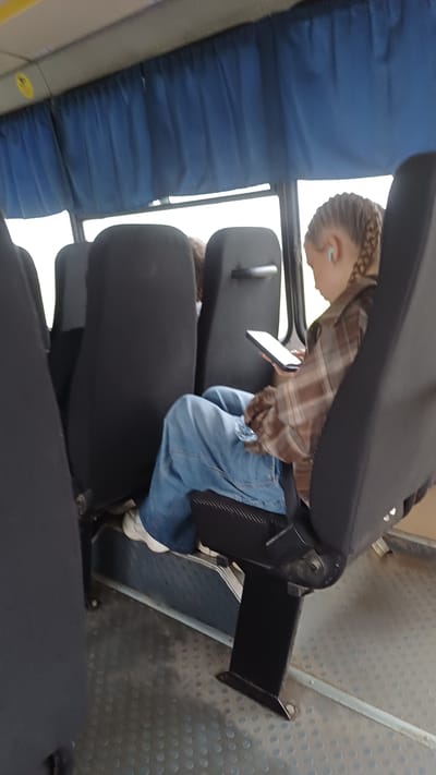 A young girl with braided hair is sitting on a bus, looking at her phone. She is wearing a brown plaid shirt and blue jeans. The bus has black seats and blue curtains. The floor of the bus is gray and textured. It is daytime, and the light outside is bright. The location is unknown, but the image may have been taken in Podgornoe, Russia.