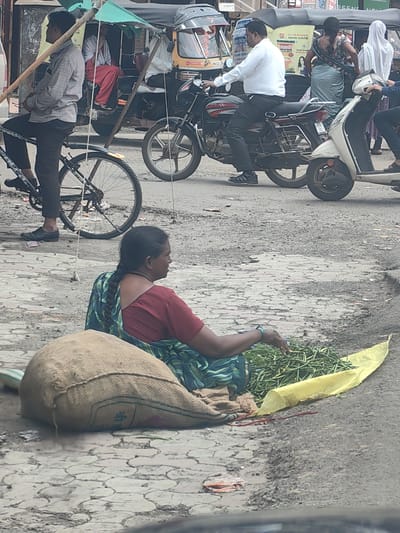 In Datala, India, on a paved road, a woman sits on the ground next to a large burlap sack and a pile of green beans spread on a yellow cloth. She is wearing a red blouse and a green and blue patterned sari, with her braided hair flowing down her back. To her right, a man in a white shirt and black pants rides a motorcycle. Behind them, an auto-rickshaw is visible, and other vehicles and people are present in the background. The scene suggests a busy roadside market or transportation hub.