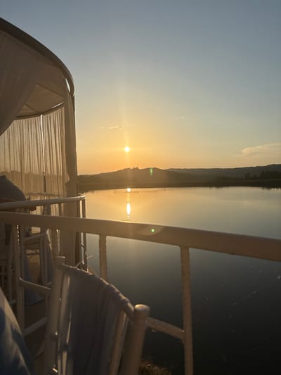 The photograph captures a serene sunset over a body of water, viewed from a covered balcony or patio. The sun, a bright orb, is low on the horizon, casting a warm, golden glow across the sky and reflecting on the calm surface of the lake. Rolling hills, densely covered in trees, form a natural backdrop to the water.
In the foreground, a white railing runs horizontally across the frame, separating the viewing area from the water. To the left, the edge of a white, curtained structure, possibly a gazebo or cabana, is visible, its light fabric gently swaying. Several white chairs are arranged on the patio, some draped with light blue fabric. The overall atmosphere is tranquil and peaceful, suggesting a moment of quiet contemplation or relaxation. The scene is located in Skalsko, Bulgaria.