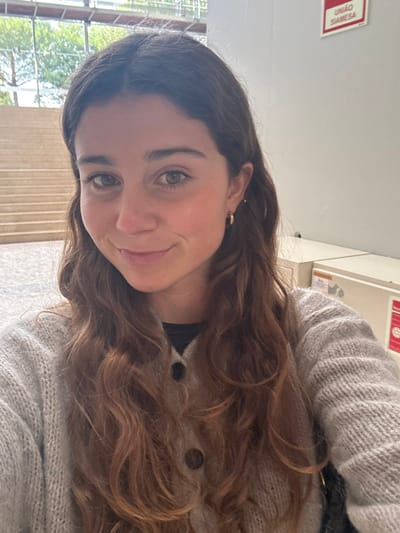 The image presents a frontal portrait of a female individual, appearing to be in her early to mid-twenties, with light skin, long wavy reddish-brown hair, and brown eyes. She exhibits a slight smile with closed lips. She is wearing small gold hoop earrings and a light grey, loosely knitted button-up cardigan over a dark undershirt.

The setting appears to be an interior or sheltered exterior space. In the background to the left, a wide, light-colored staircase ascends, and beyond it, a modern building constructed with glass and metal framing is visible, with trees partially discernible through the glass. To the right, a plain light-colored wall is present. Affixed to the upper right section of this wall is a rectangular red sign with white Portuguese text, clearly reading "UNIÃO SIAMESA" across two lines. Below this and to the subject's right, a light-colored piece of equipment or furniture featuring a partially visible red label is situated. The individual is posing for the photograph.

Based on the visible Portuguese signage ("UNIÃO SIAMESA") and the contemporary architectural elements, the scene is located within a modern building or complex in Porto Salvo, Portugal, consistent with a corporate, educational, or research park environment characteristic of the region.