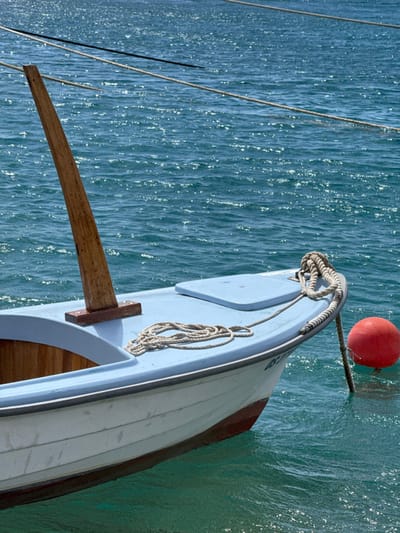 The image shows a small white boat with a wooden mast docked in the Adriatic Sea in Dugi Rat, Croatia. The boat has a light blue deck, a white hull, and a brown wooden mast that is attached to the bow. There is a white rope coiled on the deck. The boat is moored to a buoy in the water, which is visible in the lower right corner of the image. The sea is a bright blue, with light reflecting off of it. The sun is shining, and there are some small waves. The water is clear and blue, with some small waves. The scene is calm and peaceful. The coordinates, 43.446, 16.635, are consistent with a location in Dugi Rat.