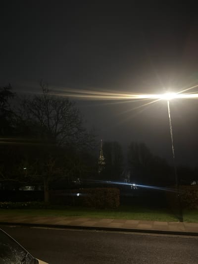 The image captures a nighttime, misty or foggy scene in Salisbury, United Kingdom, viewed from a low perspective. In the immediate foreground, the top-left section of a white vehicle's bonnet and windscreen is visible, showing water droplets. A dark, wet asphalt road surface extends across the bottom, reflecting ambient light. This road abuts a lighter-toned pavement on its right edge, which then borders a grassy verge.
In the midground, a substantial deciduous tree, bare of leaves, stands silhouetted on the left. Across the center, a manicured hedge forms a dark band. A prominent, bright street lamp on a tall pole is positioned on the right, emitting a strong, yellowish-white light that creates distinct horizontal lens flares across the image. Below the primary lens flare, a horizontal blue light streak suggests the presence of distant vehicle headlights within a darker, presumably park-like area.
The background reveals the illuminated spire of Salisbury Cathedral, partially obscured by the atmospheric conditions and surrounding dark trees. A small, distinct red light is visible at the very apex of the spire. The sky is dark and featureless, consistent with night and misty weather. No individuals are present in the scene.